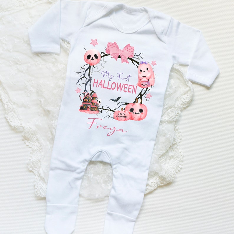 May include: White baby onesie with a pink and purple Halloween design. The design features a wreath with a skull, a ghost, a house, and pumpkins. The text "My First Halloween" and the name "Freya" are printed on the onesie.