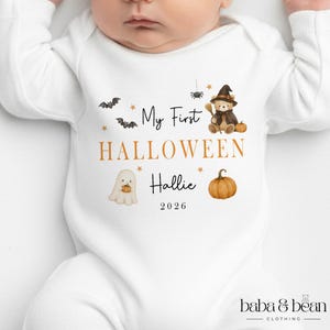Pode incluir: Um body branco de manga comprida para bebé com uma impressão gráfica com o texto My First HALLOWEEN Hallie 2026. O design inclui um urso de peluche com chapéu de bruxa, um fantasma com uma abóbora, morcegos, uma aranha e estrelas douradas.