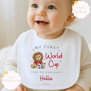 May include: A white baby bib featuring the text "MY FIRST World Cup" in red, a lion illustration, the name "Hallie", and the year "2026". The bib is made of 100% cotton. The text "COME ON ENGLAND!!!" is also visible.