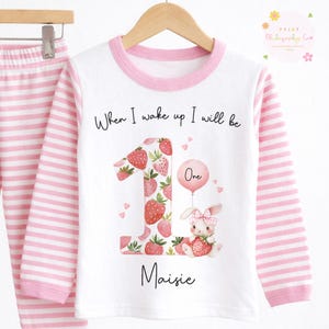 Pode incluir: Conjunto de pijama branco e rosa às riscas. A parte de cima de manga comprida tem gola e punhos rosa, com o texto "When I wake up I will be" acima de um número um grande cheio de morangos. Um coelho segura um morango.
