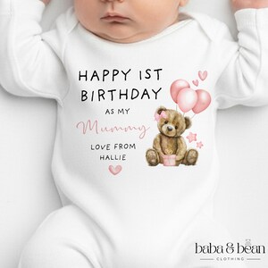 Pode incluir: Um body branco de manga comprida para beb&eacute; com uma ilustra&ccedil;&atilde;o de um urso de peluche castanho com um la&ccedil;o cor-de-rosa, bal&otilde;es cor-de-rosa e estrelas. O texto diz: HAPPY 1ST BIRTHDAY AS MY Mummy LOVE FROM HALLIE. Inclui cora&ccedil;&otilde;es e um presente.