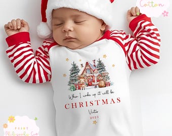 Peut inclure: Un bébé portant un pyjama de Noël à rayures rouges et blanches et un bonnet de Père Noël. Le pyjama blanc présente une scène de Noël avec le Père Noël et le texte "When I wake up it will be CHRISTMAS".