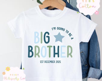 May include: White short-sleeved t-shirt with the text "I'M GOING TO BE A BIG BROTHER EST DECEMBER 2025" in blue and green. The shirt is made of 100% cotton and is hanging on a wooden hanger.