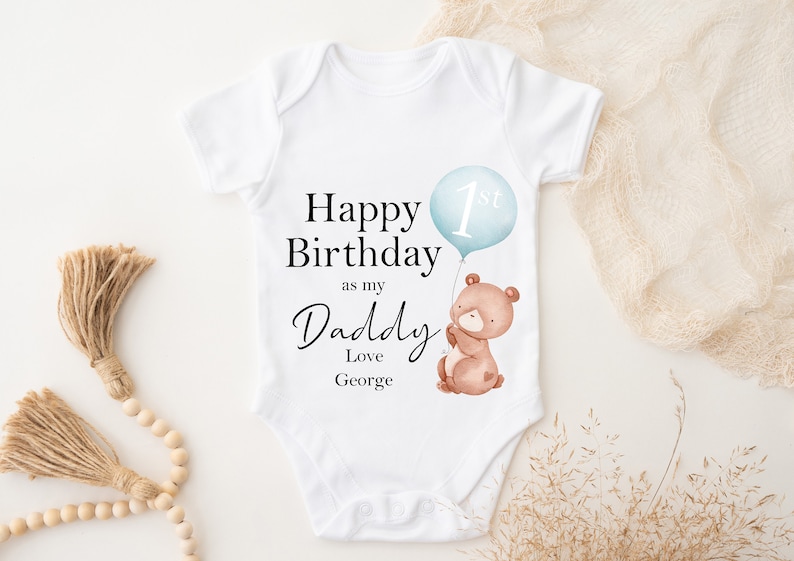 Peut inclure: Combinaison blanche pour b&eacute;b&eacute; avec le texte "Happy 1st Birthday as my Daddy Love George" et un ours en dessin anim&eacute; tenant un ballon bleu avec le num&eacute;ro 1 dessus.