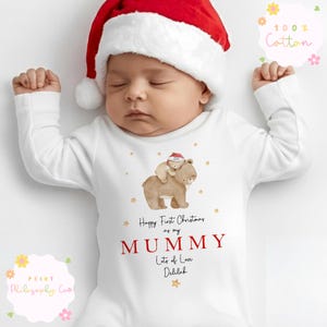 Pode incluir: Um body branco de manga comprida para bebé com uma ilustração em aguarela de um urso grande a carregar um urso pequeno com um gorro de Pai Natal. O texto diz Happy First Christmas as my MUMMY Lots of Love Delilah, rodeado por pequenas estrelas douradas.