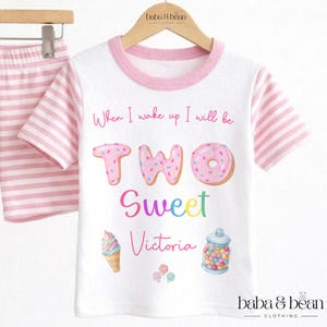 May include: A white t-shirt with pink striped sleeves and a pink collar. The shirt features the text When I wake up I will be TWO Sweet Victoria, accompanied by illustrations of a donut, an ice cream cone, a jar of candy, and lollipops.