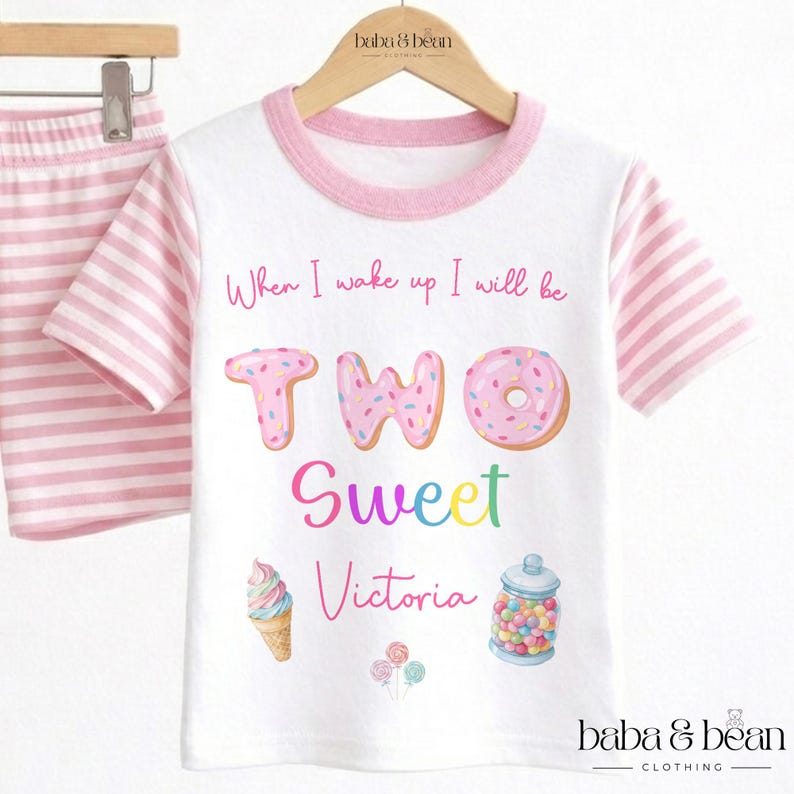 May include: A white t-shirt with pink striped sleeves and a pink collar. The shirt features the text When I wake up I will be TWO Sweet Victoria, accompanied by illustrations of a donut, an ice cream cone, a jar of candy, and lollipops.
