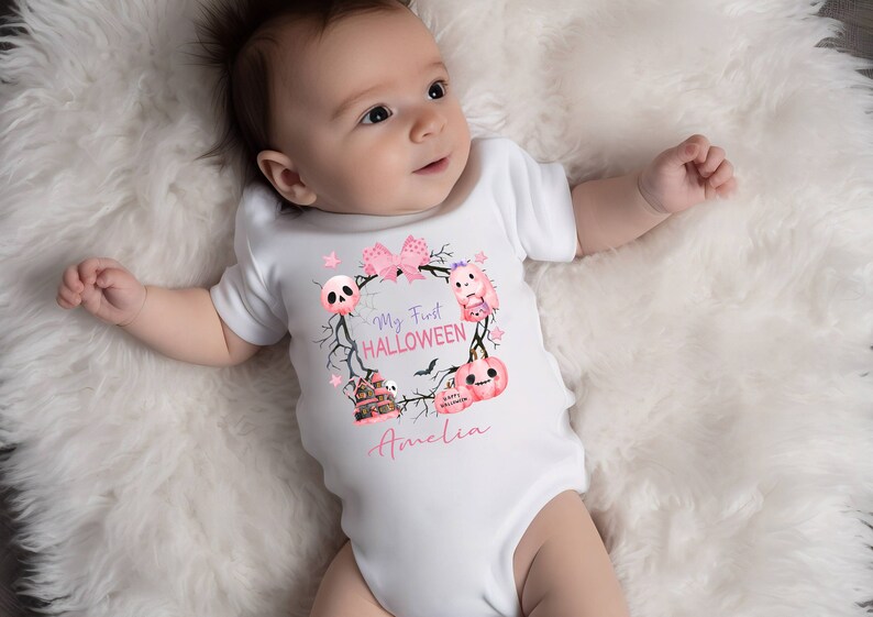 May include: A white baby onesie with a pink and purple Halloween themed design. The onesie says "My First HALLOWEEN" and features a skull, a ghost, a pumpkin, and a haunted house. The name "Amelia" is written below the design.