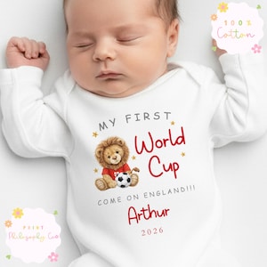 May include: A white baby bodysuit with long sleeves. It has the text "MY FIRST World Cup" in red, a lion illustration with a football, and the name "Arthur". The text "COME ON ENGLAND!!!" and the year 2026 are also printed on the bodysuit.