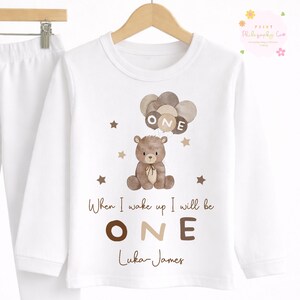 May include: White long-sleeved top featuring a brown teddy bear graphic holding balloons spelling "ONE." The text "When I wake up I will be ONE Luka-James" is also printed. A matching white pajama pant is visible.