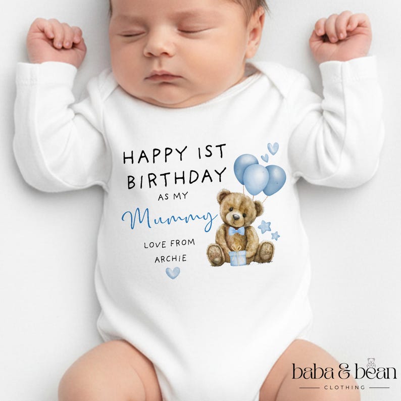 Pode incluir: Um body branco de manga comprida para beb&eacute; com uma ilustra&ccedil;&atilde;o de um urso de peluche castanho com bal&otilde;es azuis, uma gravata borboleta azul e um presente. O texto diz: HAPPY 1ST BIRTHDAY AS MY Mummy LOVE FROM ARCHIE, com cora&ccedil;&otilde;es e estrelas.