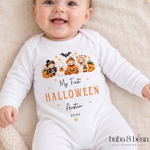 May include: A white long-sleeve baby bodysuit featuring a graphic of teddy bears in Halloween costumes, pumpkins, and a bat. The text reads My First HALLOWEEN Austin 2026, with small yellow stars scattered around the design and the brand name baba & bean.