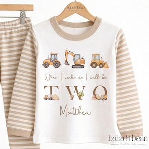 May include: A long-sleeve shirt with beige and white horizontal stripes on the sleeves and a white torso. The front features three construction vehicles, the text "When I wake up I will be TWO", and the name "Matthew" in brown lettering.
