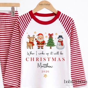 May include: A white long-sleeve pajama top with red striped sleeves and a red collar. The front features a reindeer, Santa Claus, a Christmas tree, and a snowman. Text reads: When I wake up it will be CHRISTMAS Matthew 2026.