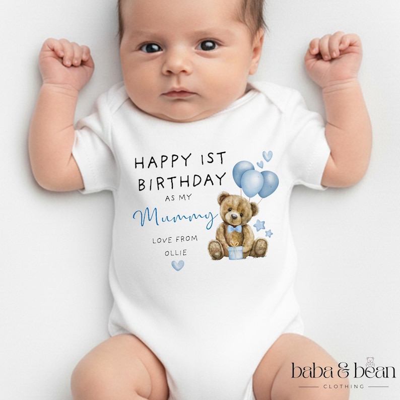 Pode incluir: Um body branco de manga curta para beb&eacute; com uma ilustra&ccedil;&atilde;o de um urso de peluche castanho com bal&otilde;es azuis e um presente. O texto diz: HAPPY 1ST BIRTHDAY AS MY Mummy LOVE FROM OLLIE. O design inclui pequenos cora&ccedil;&otilde;es e estrelas azuis.