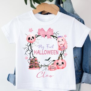 May include: White t-shirt with a pink and purple Halloween design. The design features a wreath with a bow, a ghost, a skull, a house, and a pumpkin. The text "My First HALLOWEEN" and the name "Cleo" are printed on the shirt.