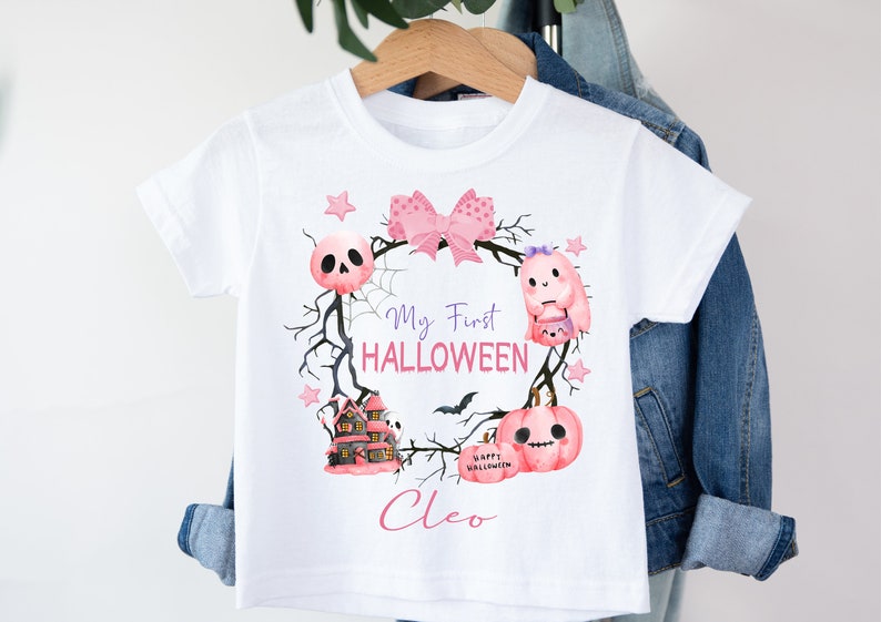 May include: White t-shirt with a pink and purple Halloween design. The design features a wreath with a bow, a ghost, a skull, a house, and a pumpkin. The text "My First HALLOWEEN" and the name "Cleo" are printed on the shirt.