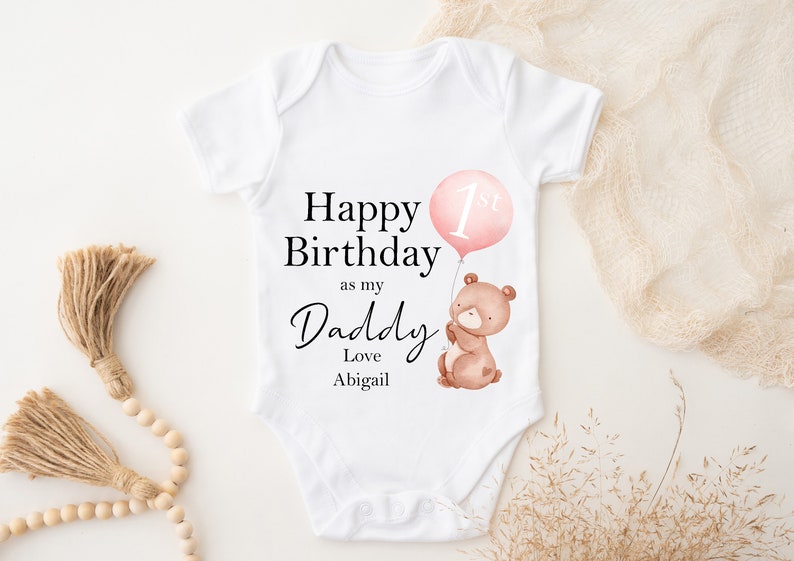 Peut inclure: Combinaison blanche pour b&eacute;b&eacute; avec le texte "Happy Birthday as my Daddy Love Abigail" et un ballon rose avec le chiffre 1 dessus. Un ours brun avec un nez rose tient le ballon.