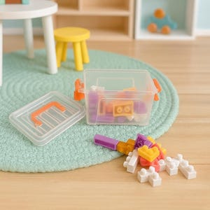 Könnte beinhalten: Eine transparente Kunststoff-Aufbewahrungsbox mit orangefarbenem Griff und Verschluss, gefüllt mit bunten Spielzeugbausteinen. Der Deckel ist offen, und einige Blöcke sind in der Nähe verstreut. Die Szene umfasst einen kleinen Tisch, einen Hocker und ein holzförmiges Regal.