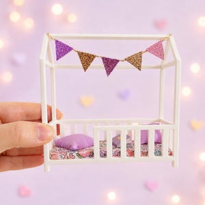 Könnte beinhalten: Miniatur-Bettrahmen in Weiß mit floral gemusterter Matratze, lila Kissen und Girlande aus glitzernden Dreiecksfahnen in Gold, Rosa und Lila. Ein Puppenhaus-Zubehör.