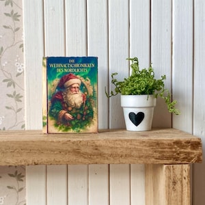 Könnte beinhalten: Ein Miniaturbuch mit dem Titel "Die Weihnachtschroniken des Nordlichts" mit dem Weihnachtsmann, neben einem weißen Topf mit schwarzem Herz, der eine grüne Pflanze enthält. Die Gegenstände stehen auf einem Holzregal.