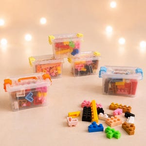 Op de afbeelding: Vier doorzichtige plastic opbergdozen met gekleurde sluitingen en handvatten, elk gevuld met diverse kleine bouwstenen. Een verzameling losse, veelkleurige plastic bouwstenen ligt verspreid op de voorgrond.