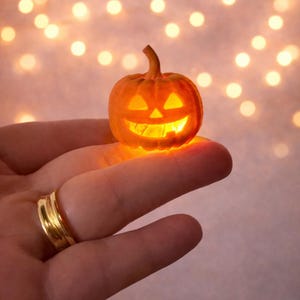 Può includere: Una piccola zucca arancione scolpita con un volto illuminato, tenuta in una mano. La zucca ha un gambo e una faccia sorridente di Jack-o'-lantern. Lo sfondo presenta luci morbide e sfocate dai toni caldi, creando un tema festivo di Halloween.