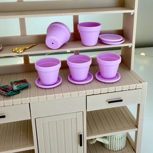 Puede incluir: Un banco de macetas en miniatura con varias macetas y platillos de color lavanda. El banco tiene estantes, cajones y un armario. También se ven unas tijeras doradas y pequeños paquetes. La combinación de colores general es un beige suave y neutro.