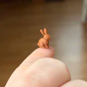 Może przedstawiać: Mała, pomarańczowo-brązowa figurka królika. Miniaturowy królik siedzi na czubku palca, prezentując swoje wyprostowane uszy i szczegółowe cechy. Figurka to uroczy przedmiot dekoracyjny.