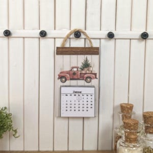 Könnte beinhalten: Ein Miniatur-Holzkalender mit rustikalem Design. Der Kalender zeigt einen roten LKW, der einen Weihnachtsbaum und Geschenke transportiert. Der Monat Januar wird angezeigt. Der Kalender hängt an einer weißen Holzwand mit schwarzen Knöpfen.