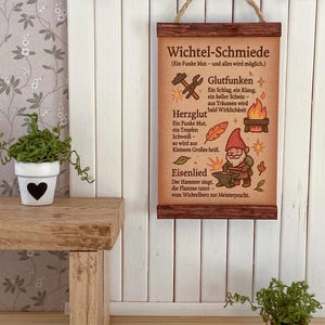 Op de afbeelding: Een klein houten bord met de tekst "Wichtel-Schmiede" en illustraties van gereedschap, een vuur en een kabouter. Het bord is bruin met een touwhanger en wordt weergegeven tegen een witte wand met panelen.