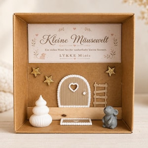 Könnte beinhalten: Eine braune Pappschachtel mit einem Miniatur-Set mit der Aufschrift Kleine Mäusewelt und LYKKE Minis. Es enthält eine beige Bogentür mit Herzausschnitt, eine kleine Leiter, drei goldene Glitzersterne, eine weiße Tannenfigur und eine graue Maus.