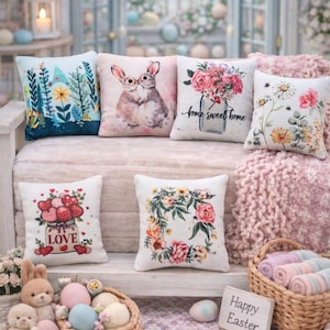 Peut inclure: Plusieurs coussins décoratifs avec des motifs floraux et de lapins. Les coussins sont disposés sur une surface rose clair, avec un panier d'œufs de Pâques et une pancarte indiquant "Happy Easter".