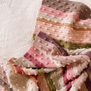 Puede incluir: Una manta de crochet multicolor con un patrón de punto de relieve. La manta presenta rayas de color rosa, marrón, beige y verde.