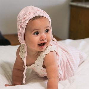 Puede incluir: Un bebé con un gorro de crochet rosa con ribete blanco y un vestido a rayas rosa y blanco. El bebé está gateando sobre una superficie blanca.
