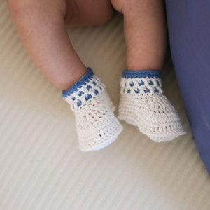 May include: A pair of white crocheted baby booties with blue trim. The booties are on a white surface.