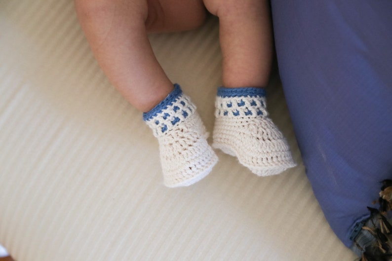 May include: A pair of white crocheted baby booties with blue trim. The booties are on a white surface.