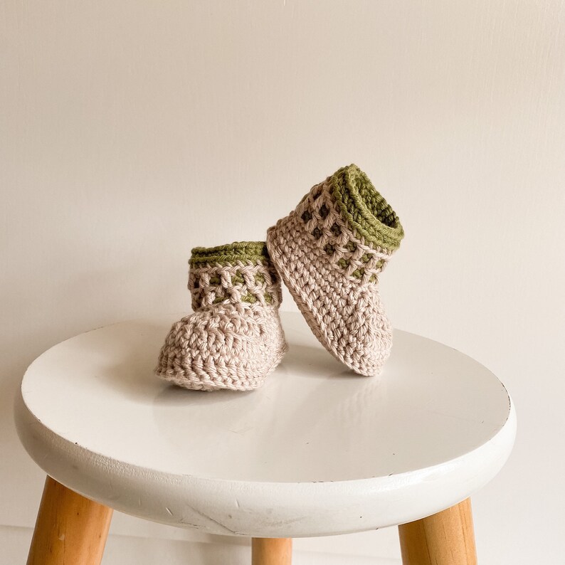 May include: A pair of crocheted baby booties in beige with green trim. The booties are sitting on a white stool.