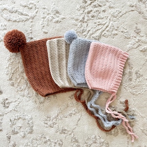 May include: Four crocheted baby hats in different colors: brown, beige, gray, and pink. Each hat has a pom-pom on top and ties under the chin.