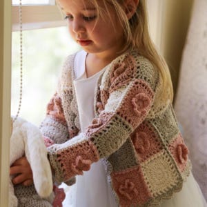 Puede incluir: Un cárdigan de ganchillo hecho a mano para niños en colores pastel, incluyendo rosa, crema y gris. El cárdigan presenta un diseño floral y lo lleva una niña pequeña. El trabajo de ganchillo es detallado, con puntadas visibles y patrones texturizados, creando una estética encantadora.