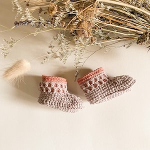 May include: A pair of crocheted baby booties in a light brown color with a pink trim. The booties have a textured pattern.