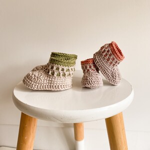 May include: Two pairs of crocheted baby booties. One pair is beige with a green crocheted cuff, and the other pair is beige with a pink crocheted cuff. The booties are sitting on a white stool with wooden legs.
