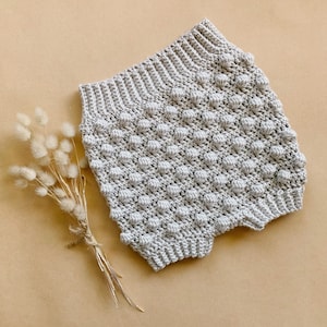 May include: A pair of beige crocheted baby bloomers with a textured, bobble stitch pattern.