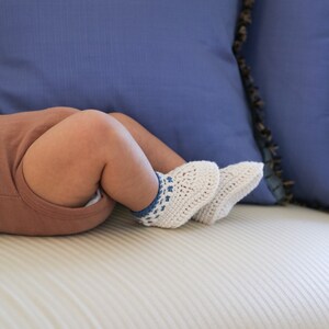 May include: A baby wearing brown pants and white crocheted booties with blue accents, lying on a white couch with blue pillows.