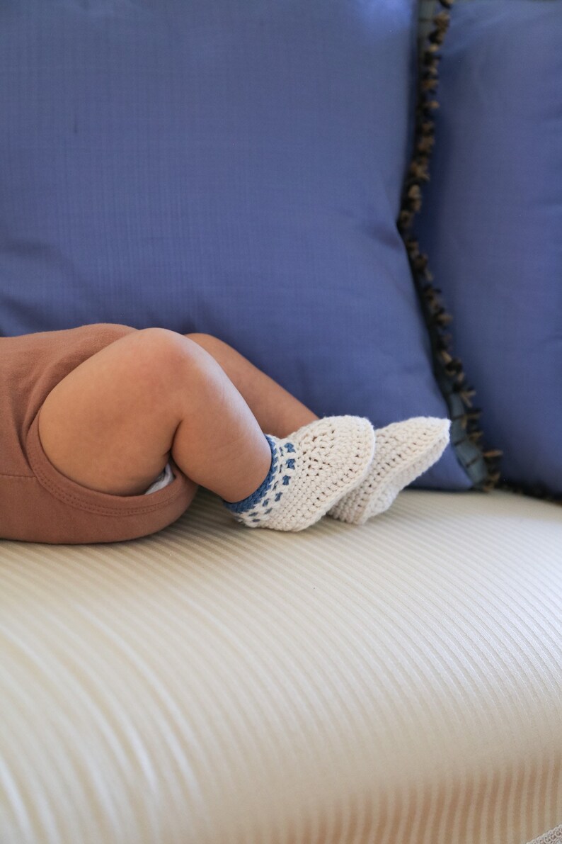 May include: A baby wearing brown pants and white crocheted booties with blue accents, lying on a white couch with blue pillows.