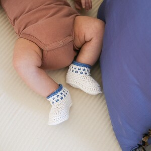 May include: A baby wearing a brown onesie and white crocheted booties with blue trim. The booties have a white crocheted pattern and blue trim.