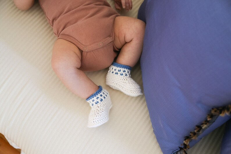 May include: A baby wearing a brown onesie and white crocheted booties with blue trim. The booties have a white crocheted pattern and blue trim.