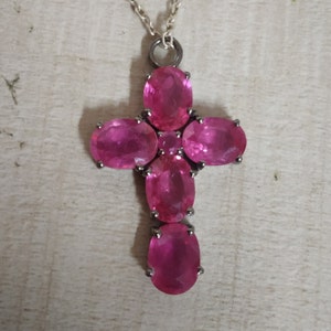 May include: A silver chain necklace with a pink gemstone cross pendant. The cross is made up of five oval-shaped pink gemstones.