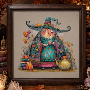 May include: A framed cross-stitch artwork depicting a witch wearing a wide-brimmed teal hat and a patterned cloak, holding a small cup. The scene includes a stack of books, a pumpkin, and a golden teapot, all set against a neutral background.