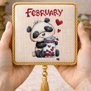 以下が含まれることがあります： 赤いハートでいっぱいの瓶を持っているパンダが描かれたクロスステッチ。上部には「February」の文字。正方形は金色のトリムと金色のタッセルで縁取られています。